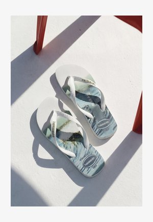 Paar weiße Havaianas Flip-Flops mit blau-grauem Wellenmuster, auf weißer Oberfläche unter einem roten Stuhl, der Schatten wirft.