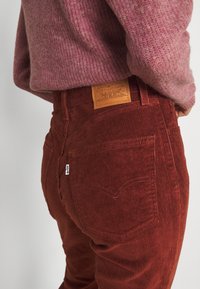 Pantalon en velours côtelé de couleur rouille avec des rayures verticales, doté d'une étiquette en cuir à l'arrière de la ceinture et d'une petite étiquette logo sur le côté.