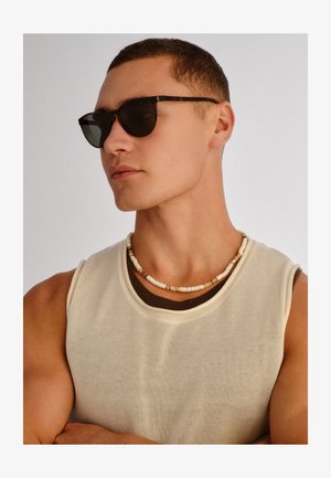 Jeune homme aux cheveux courts portant des lunettes de soleil foncées, un haut beige sans manches, un collier superposé, regardant vers la gauche sur un fond uni.