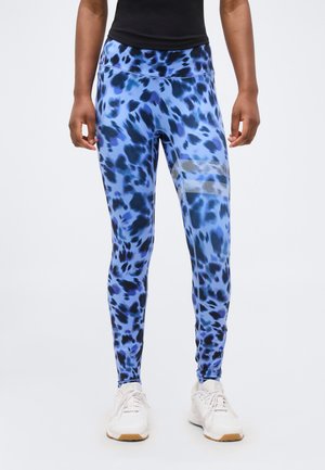 Osoba oblečená v modro-čiernych legínach s bielymi bodkovanými pruhmi na stehne, v bielych teniskách a čiernom topu, stojaca na bielom pozadí.