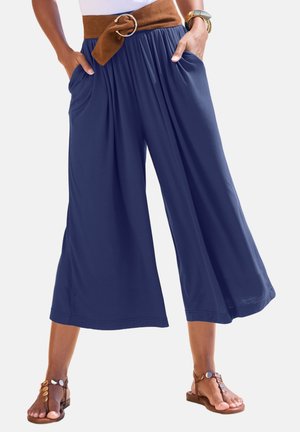 Woman wearing navy wide-leg cropped pants with side pockets, a brown belt, and brown sandals, standing with hands in pockets.