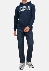 Dunkelblauer Kapuzenpullover mit weißem "JACK & JONES"-Schriftzug, Kängurutasche, gerippte Bündchen und Saum, kombiniert mit dunklen Jeans und Turnschuhen.