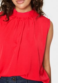 Blusa rossa senza maniche con tessuto plissettato e design a collo alto, caratterizzata da una texture liscia e una silhouette drappeggiata. Indossata sopra jeans scuri.