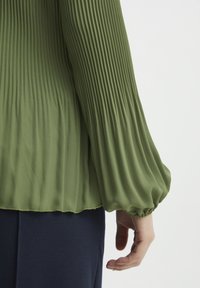 Blusa verde plissettata con maniche lunghe e arricciate e una texture morbida, indossata sopra un indumento di un blu scuro. Tessuto liscio e design fluido.
