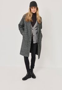 Grauer gemusterter Mantel mit großen Taschen, über einem grauen Cardigan und einem dunklen Rollkragenpullover getragen. Kombiniert mit schwarzen Jeans und Ankle-Boots. Schwarze Kappe.