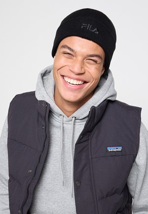 Joven sonriente, con un gorro negro de FILA, sudadera gris y chaleco negro de Patagonia, frente a un fondo claro y liso.
