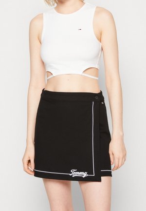 Nainen pukeutuneena valkoiseen lyhyeen toppiin ja mustaan minihameeseen, jossa on valkoinen reunus ja helmassa kirjailtu "Tommy"-teksti.