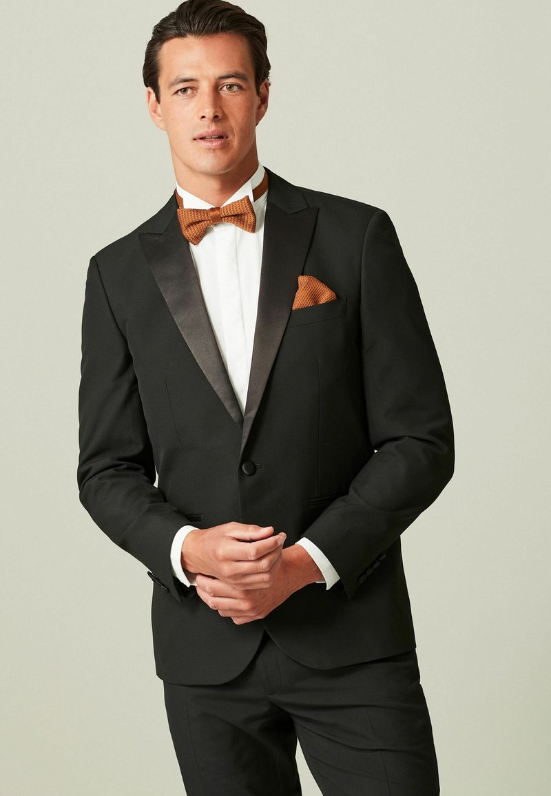 Tuxedo negru cu rever din satin, asortat cu o cămașă albă și o cravată fluture portocalie. O batista texturată completează ansamblul.