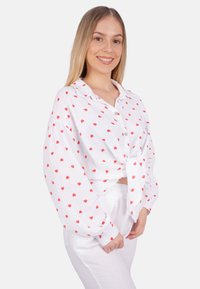 White shirt with red heart pattern, button-down design, long sleeves, knotted at the waist, made from textured fabric. Worn with white pants.