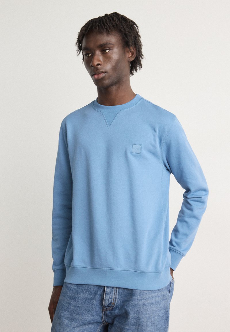 Sudadera azul claro con cuello redondo, puños y dobladillo acanalados. Presenta un pequeño logo en relieve en la zona izquierda del pecho. Textura suave.