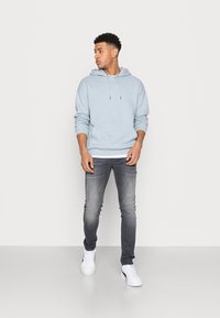 Lyseblå hettegenser med snøring og kengurulomme, kombinert med mørk grå jeans og hvite sneakers, mot en ensfarget hvit bakgrunn.