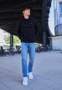 Felpa nera con cappuccio e tasca frontale, jeans blu chiaro e sneakers bianche. Sullo sfondo si vedono biciclette e un ambiente architettonico moderno.