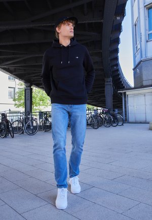 Svart huva sweatshirt med framficka, ljusblå jeans och vita sneakers. Bakgrunden visar cyklar och en modern arkitektonisk miljö.
