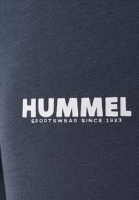 Zbliżenie na ciemną tkaninę z nadrukiem białego tekstu „HUMMEL SPORTSWEAR SINCE 1923”.