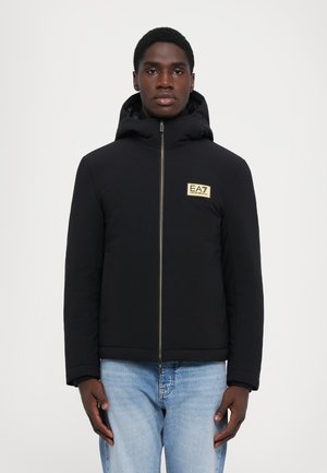 Black zip-up jacket with a hood, featuring a gold logo patch, textured fabric, straight cut, and concealed side pockets.