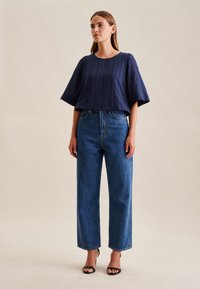 Top corto a maniche corte in blu navy con texture abbinato a jeans blu a vita alta e di taglio largo, indossato con sandali neri con tacco, in piedi davanti a uno sfondo neutro.