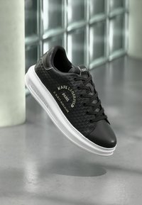 Black leather sneakers with a textured side panel, white sole, black laces, and gold branding reading "Karl Lagerfeld Paris."