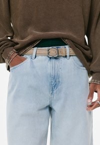 Barna textúrájú, hosszú ujjú ing világoskék farmernadrággal párosítva. Bézs öv ezüst csattal. Kezek a zsebekben.