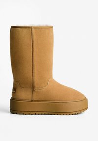 Stivale al ginocchio in suede beige con una morbida fodera interna, suola in gomma resistente e accenti di cucitura minimal, caratterizzato da un piccolo tag con il marchio sul lato.