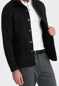 Homme portant une veste noire boutonnée sur un polo blanc et un pantalon gris, tenant la veste ouverte d'une main.
