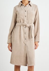 Abito camicia in corduroy beige con maniche lunghe, colletto strutturato, chiusura frontale a bottoni, due tasche sul petto e cintura abbinata da legare.