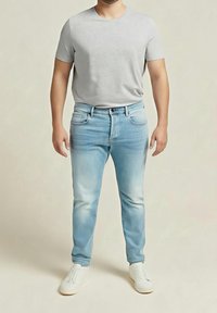 Homme portant un t-shirt gris clair, un jean bleu clair et des baskets blanches, debout devant un fond beige uni.