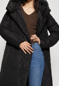 Kvinna i en svart puffjacka över en brun stickad V-ringad tröja och blå denimjeans, som justerar framdelen av jackan med händerna.