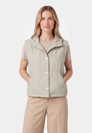 Lyshåret kvinde iført en beige kortærmet skjorte, lysebeige ærmeløs knapvest med lommer og beige bukser, stående med hænderne i lommerne.
