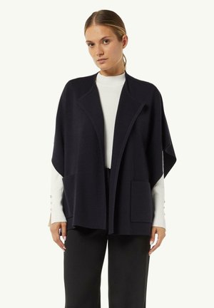 Übergroßer Cardigan aus einer Wollmischung in Marineblau mit einem Schalkragen, zwei vorderen Taschen und lockerem Schnitt. Kombiniert mit einem weißen gerippten Pullover.