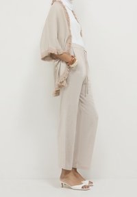 Beżowe kimono z otwartym przodem z frędzlami, zestawione z jasnobeżowymi spodniami o szerokich nogawkach i białymi sandałami typu mule z odkrytymi palcami. Gładka tekstura na całej powierzchni.