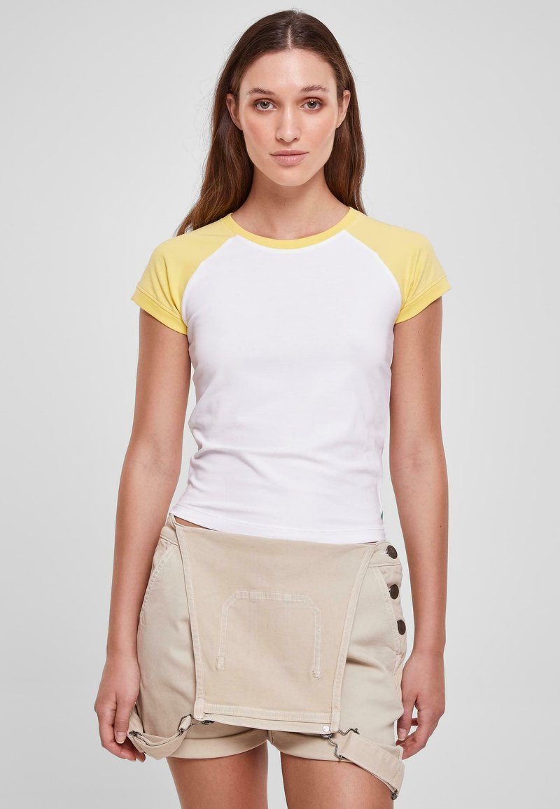 Hvit t-skjorte med korte ermer og gule raglan-ermer, kombinert med beige shorts med lommer og trykknapper for lukking.