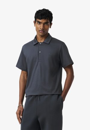 Joven con camisa polo de manga corta gris oscuro y pantalones a juego, de pie con una mano en el bolsillo contra un fondo blanco.