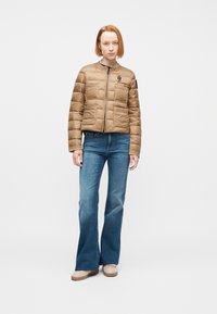 Γυναίκα με κοντά κόκκινα μαλλιά, που φοράει ταμπά μπουφάν με ραφές, μπλε τζιν με φαρδιά μπατζάκια και μπεζ παπούτσια, που στέκεται μπροστά σε ένα λευκό φόντο.