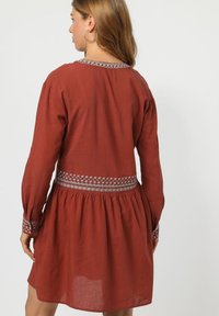 Vestido de color óxido con mangas largas, escote y puños bordados, cintura fruncida y falda acampanada, hecho de una tela texturizada.