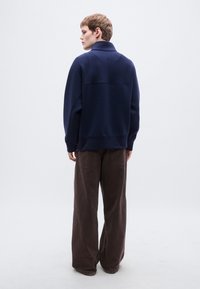 Πουλόβερ σε ναυτικό μπλε με ψηλό γιακά και ραφή στον ώμο, συνδυασμένο με φαρδιά καφέ παντελόνια από υφή υφάσματος.
