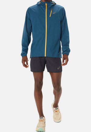 Mann trägt eine türkisfarbene Kapuzenjacke mit gelbem Reißverschluss, schwarze Shorts und beige Laufschuhe, steht vor einem einfarbigen Hintergrund.