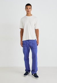 Světle béžové bavlněné tričko, volný střih, doplněné modrými kordovým cargo kalhotami s kapsami. Černé tenisky dotvářejí outfit.