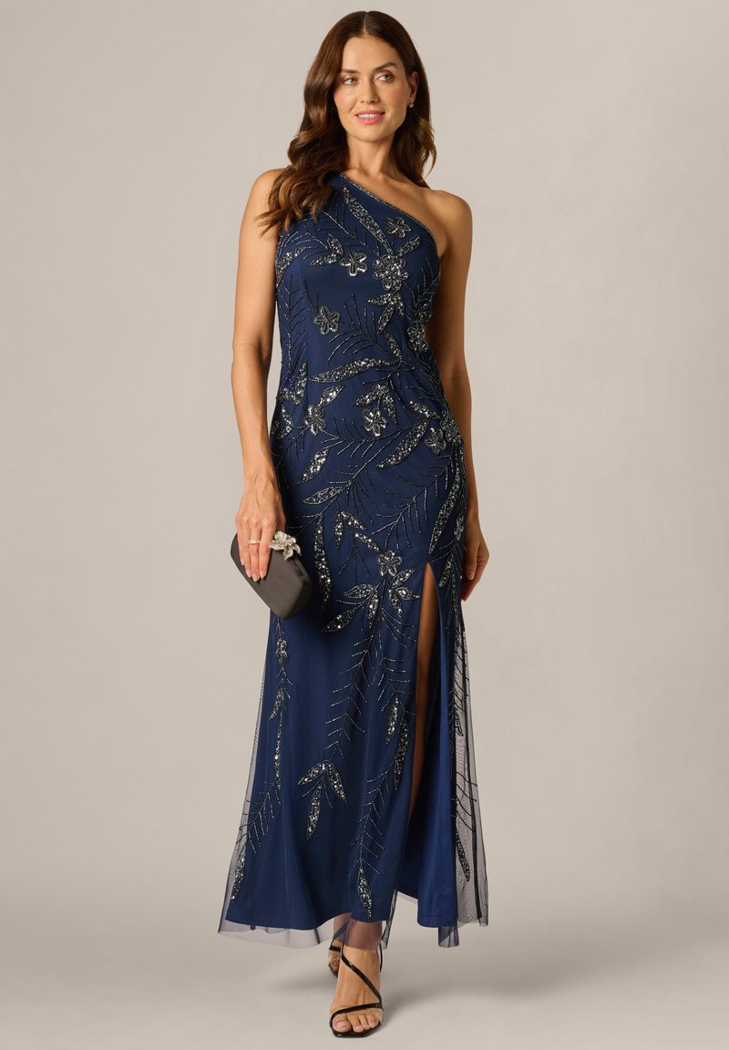 Kvinde iført navyblå aftenkjole med én skulder udsmykket med sølvfarvede blomsterformede palietter, med slids ved låret, sorte clutch og sorte sandaler med stropper, stående foran en beige baggrund.