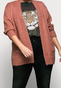 Rose-colored knitted cardigan with ribbed edges, open front, and two pockets, worn over a graphic tiger print T-shirt and black pants.