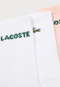 Δύο ζευγάρια βαμβακερές κάλτσες: μία λευκή με πράσινο κείμενο "LACOSTE" και λογότυπο κροκόδειλου, και μία ανοιχτό ροζ με αντίστοιχο πράσινο λογότυπο.