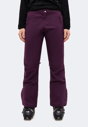 Une personne porte un pantalon de ski couleur prune avec des poches zippées et des chaussures de ski noires, sur un fond clair uni.