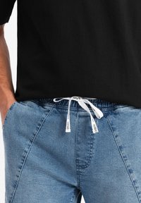 Pantalones de mezclilla en azul claro con cintura elástica y cordón blanco. Presentan un diseño de cinco bolsillos y una textura sutil. Camiseta negra arriba.