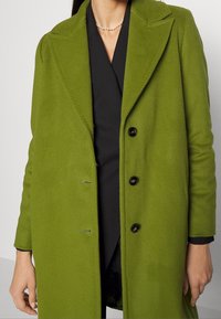 Grüner Wollmantel mit Reverskragen, sechs Knöpfen und glatter Textur. Wird über einem schwarzen Oberteil getragen und zeigt ein maßgeschneidertes Design.