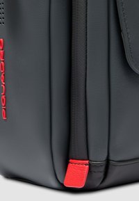 Zwarte leren tas met rode accenten, met een gladde textuur, subtiele stiksels en een rechthoekige vorm. Piquadro-logo zichtbaar.