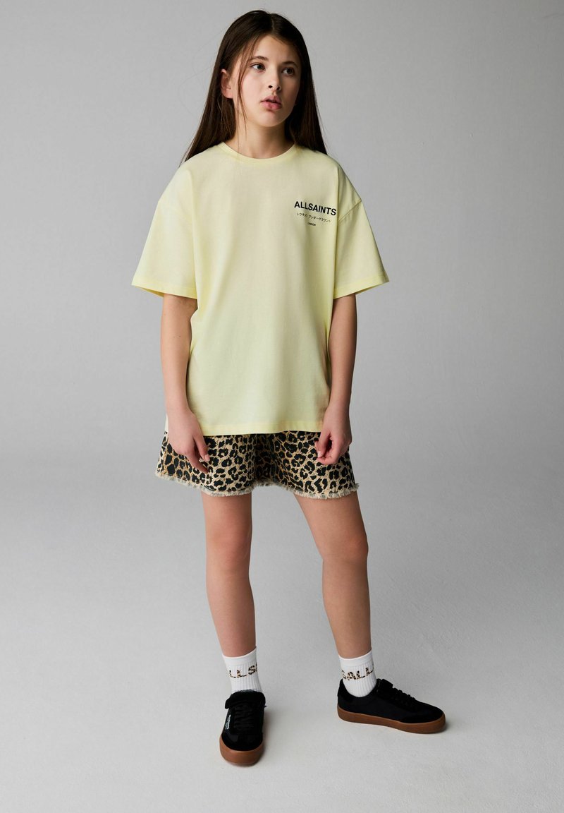 Mladé dievča stojí oblečené v svetložltom tričku AllSaints, leopardích kraťasoch, bielych ponožkách a čiernych teniskách na šedom pozadí.