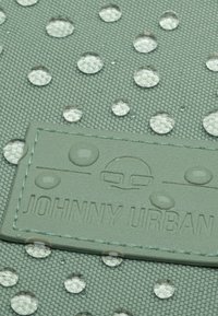 Zöld textúrált szövet tiszta vízcseppekkel és egy gumírozott címkével, amelyen az "JOHNNY URBAN" felirat található, minimalista dizájnnal.