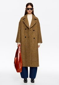Abrigo de lana marrón en patrón de espiga con un cuello ancho, diseño de doble botonadura, combinado con jeans de pierna ancha azul oscuro y un bolso de cuero rojo.