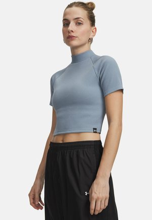 Vrouw met haar in een knot, draag een lichtblauw kort mouwlang croptop en zwarte elastische taillebroek, staand tegen een effen achtergrond.
