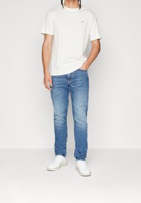 Vit t-shirt med kort ärm och logotyp, blå slim-fit jeans och vita sneakers, med en enkel och avslappnad design.