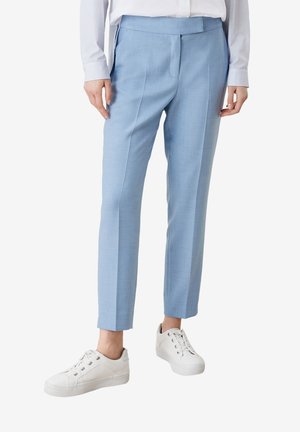 Persona vistiendo pantalones a medida azul claro, camisa blanca abotonada y zapatillas blancas, de pie contra un fondo liso.